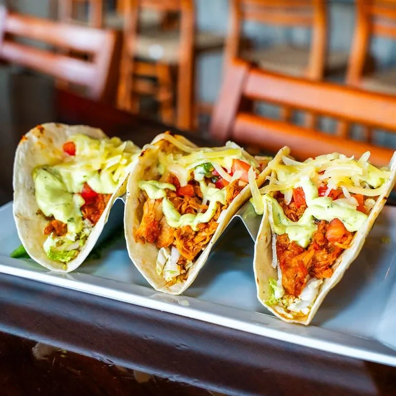 Three soft-shell chicken tacos with cheese, vegetables, and green sauce.