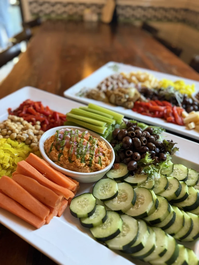 Two white platters filled with hummus, fresh vegetables, olives, and chickpeas.