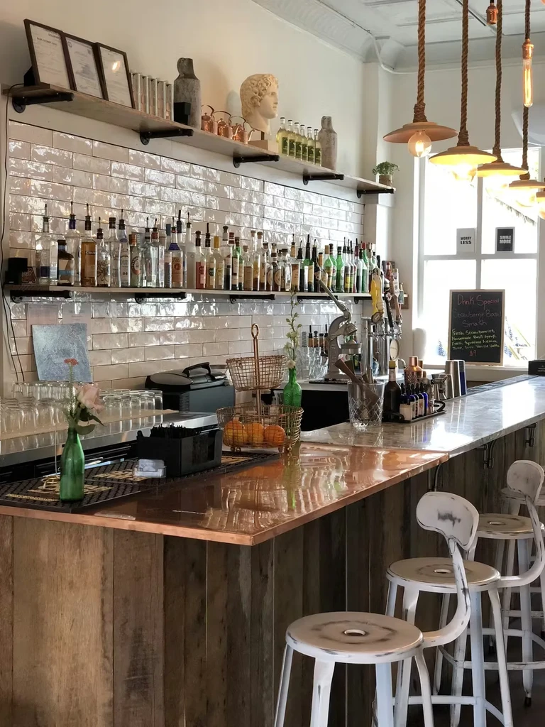 Empty bar with copper-top counter, spirit bottles, and distressed metal stools.