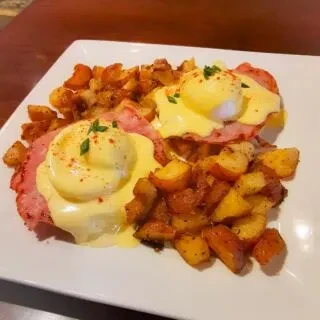 Two Eggs Benedict topped with hollandaise, beside roasted potatoes.