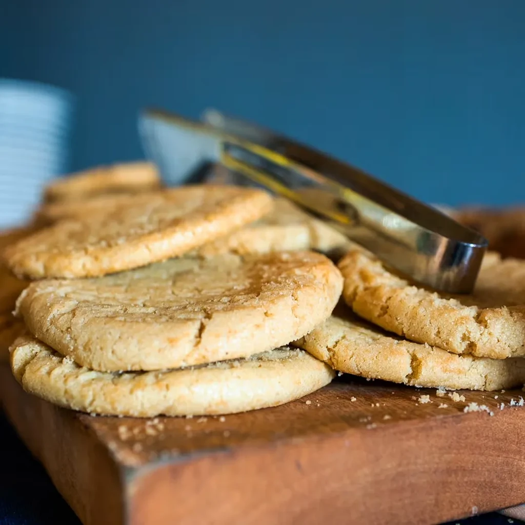 Light brown cookies stacked on a rustic wooden board with tongs.
