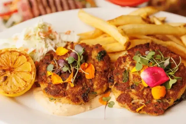 Plated crab cakes garnished with microgreens, edible flowers, lemon, fries, coleslaw.
