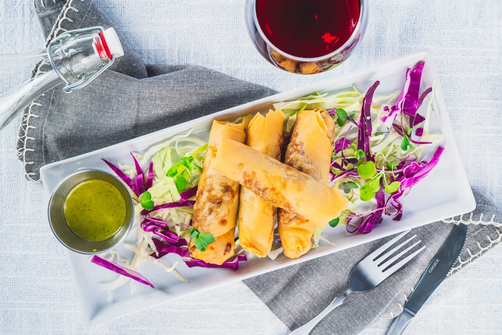Overhead shot of six golden-brown fried rolls, green sauce, wine, and cutlery.