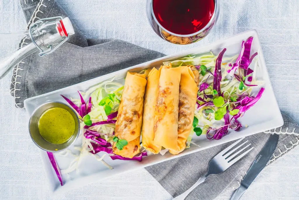 Golden spring rolls on white plate with cabbage, green sauce, and drink.