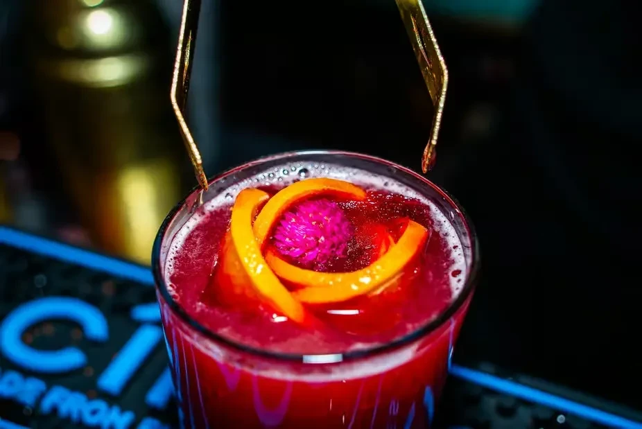 Close-up red cocktail in glass, garnished with orange peel and flower.