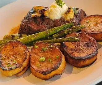 Plated steak with creamy topping, asparagus, roasted potatoes, and herbs.