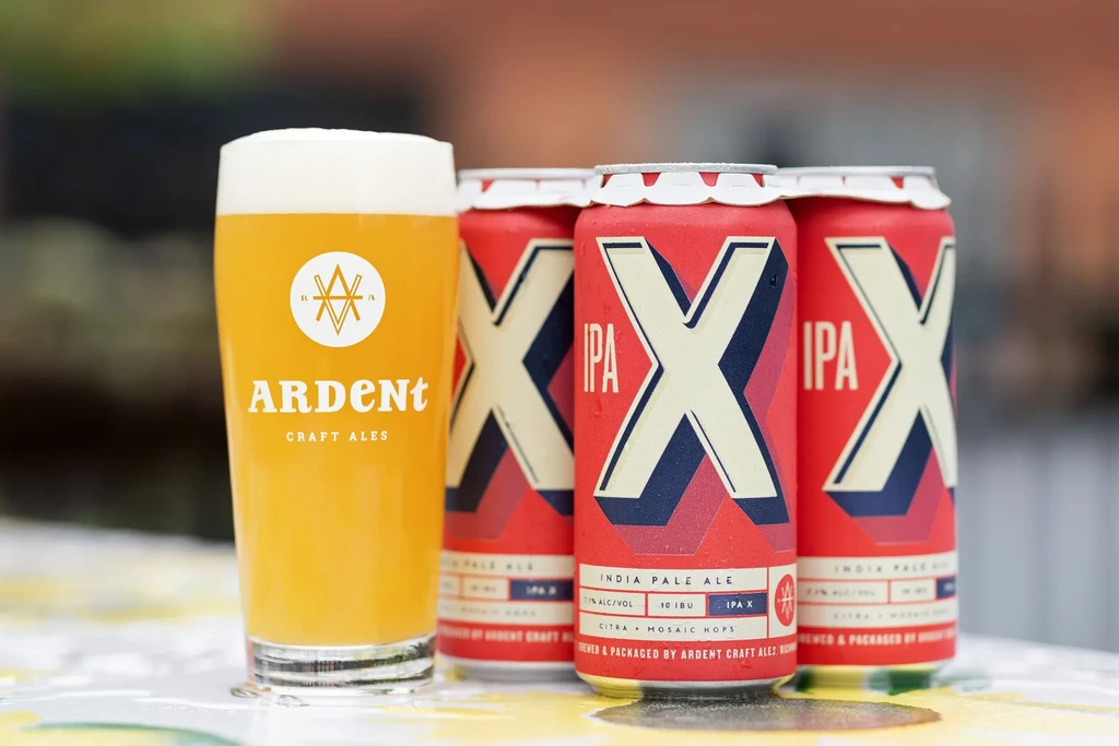 A glass of cloudy Ardent Craft Ales beer with three red IPA cans.