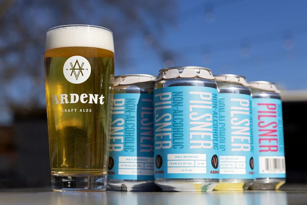 Glass of Ardent non-alcoholic pilsner with stacked cans.