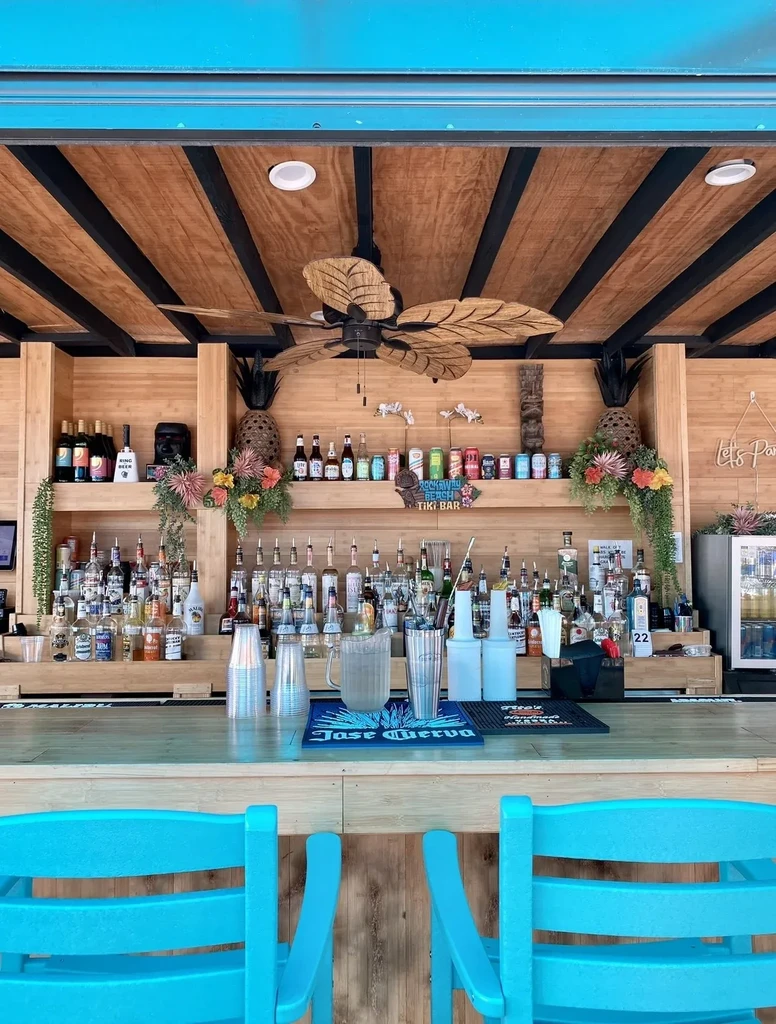 Vibrant tiki bar with liquor bottles, turquoise stools, Rockaway Beach sign.