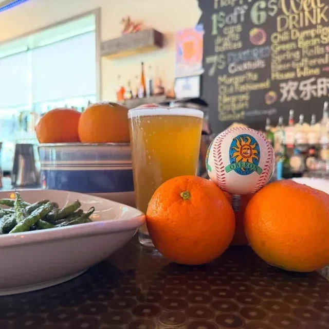 Beer, green vegetables, oranges, and an Oberon baseball on a bar counter.