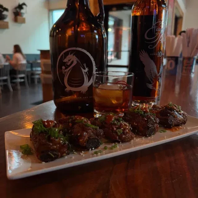 Glazed ribs, whiskey with ice sphere, and two bottles on a dark bar.