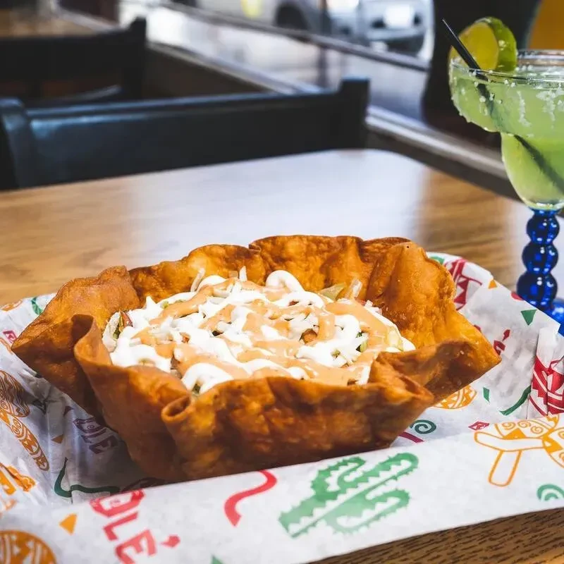 Close-up: Taco salad in fried bowl, sauces, margarita, casual restaurant.