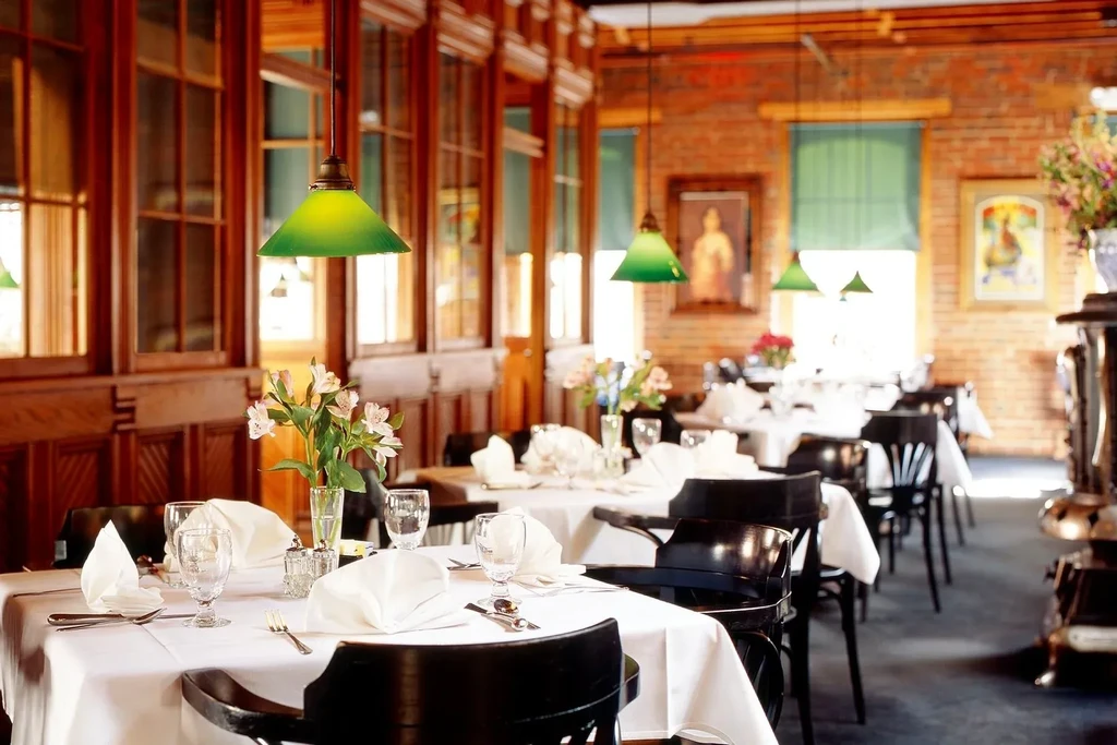 Restaurant dining room with set tables, white linens, flowers, wood, brick.