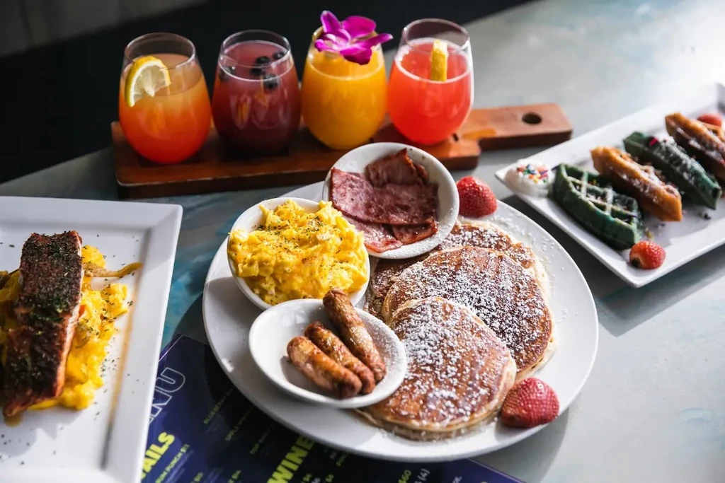 Vibrant brunch spread with salmon, pancakes, bacon, eggs, waffles, and colorful drinks.