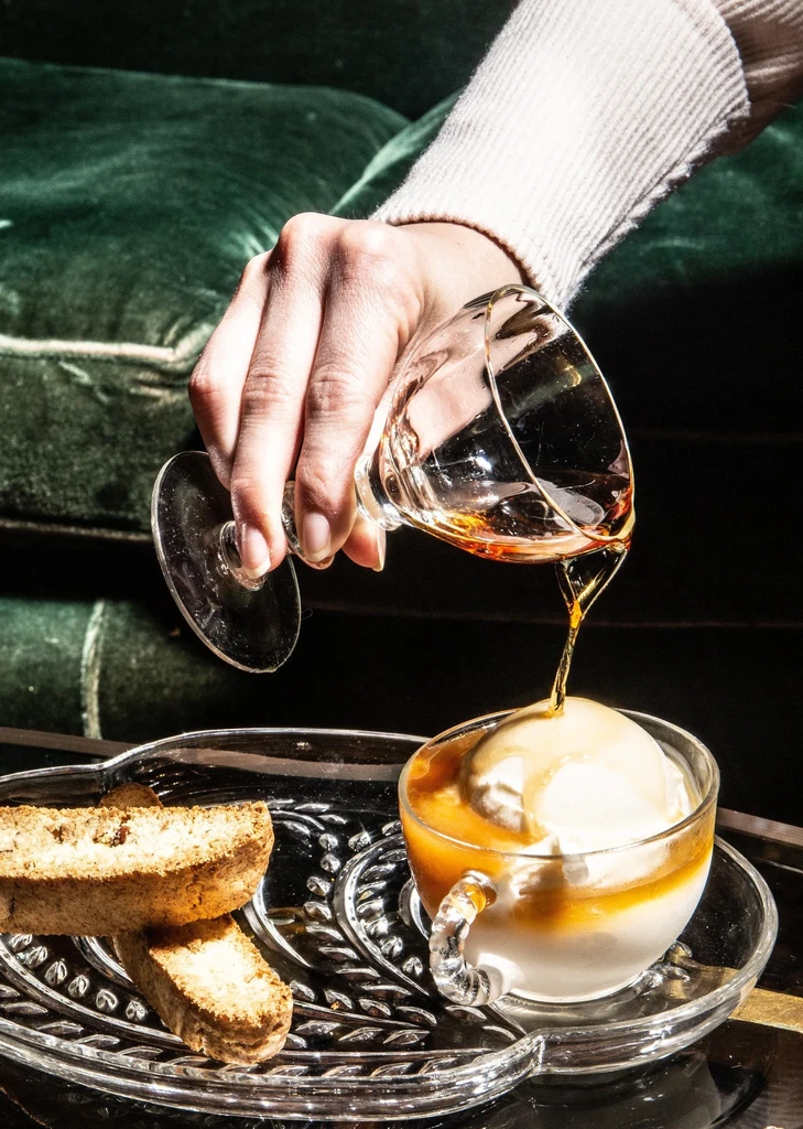 Hand pours golden liquid over vanilla ice cream, served with biscotti.