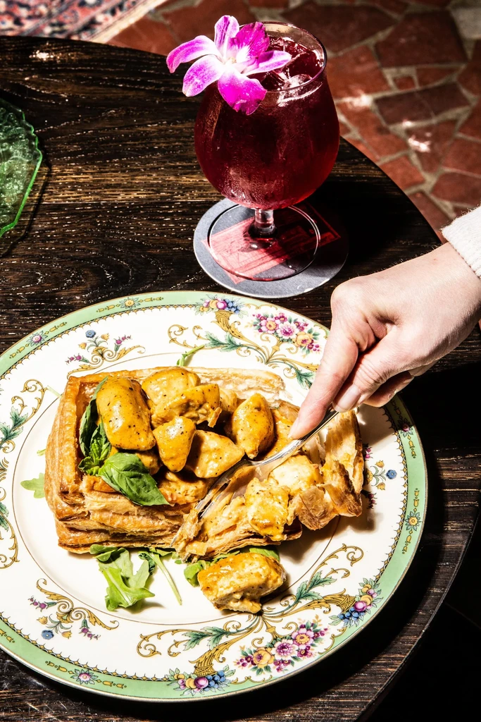 Hand forks creamy chicken pastry next to a purple orchid drink.