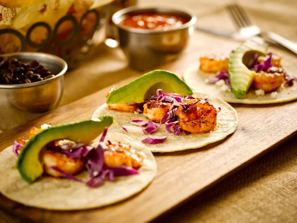 Three shrimp tacos with purple cabbage and avocado on a wooden board.