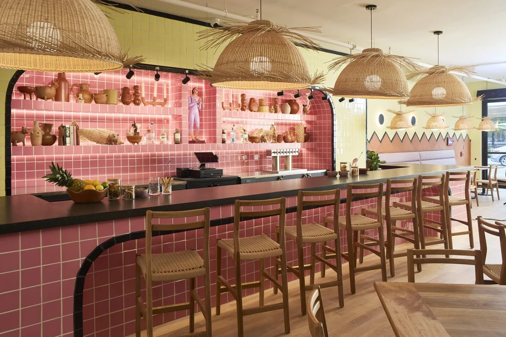 Modern bar interior with pink-tiled counter, fruit, and woven pendant lights.