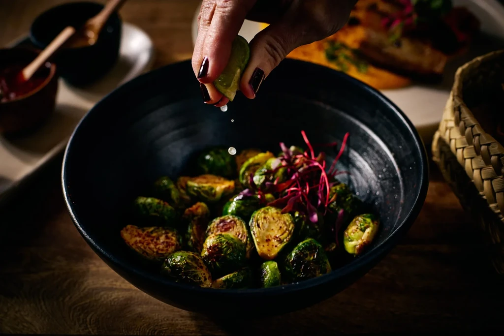 Hand squeezing lime over roasted brussels sprouts in a dark bowl.