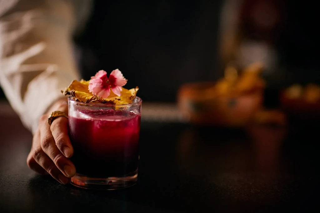Dark red cocktail garnished with dried pineapple and pink flower, held by a hand.