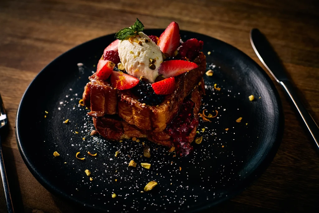 Stacked French toast with berries, cream, and pistachios on a black plate.