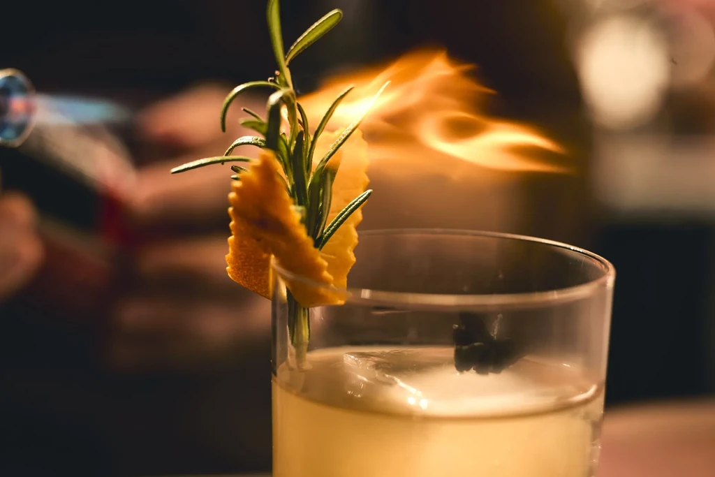 Close-up of a hand torching a cocktail with a flaming rosemary garnish.