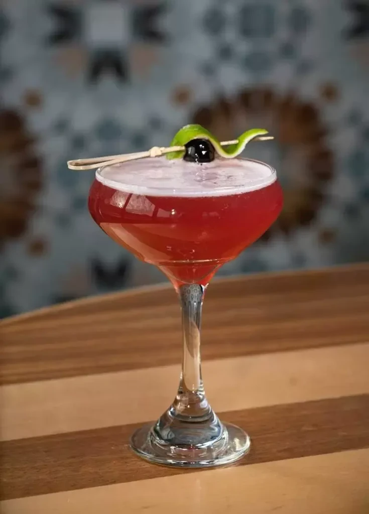 Red cocktail in coupe with frothy head, cherry, lime swirl.