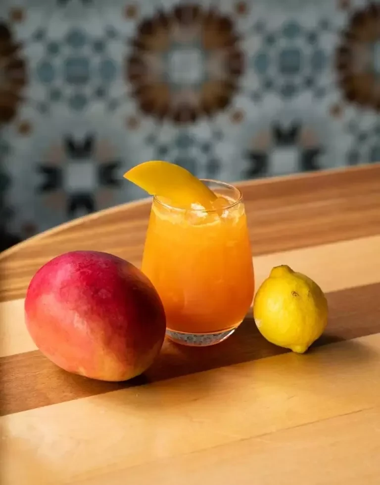 Orange drink with mango slice, whole mango, and lemon on wood.