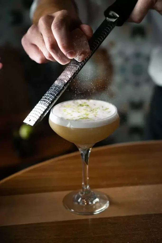 Hands zesting citrus onto a frothy cocktail in a coupe glass.