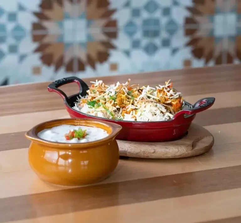 Garnished biryani in red cast-iron pan and raita on wood table.