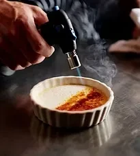 Hand holding kitchen torch caramelizes crème brûlée; smoke rises.