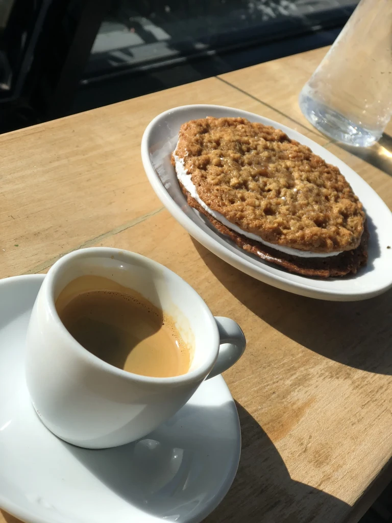 Espresso, oatmeal cream pie, and water on a sunlit wooden table.