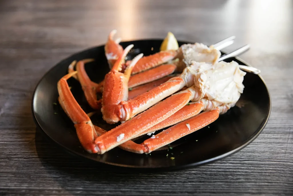 Cooked orange-red snow crab legs, lemon, crab tools on black plate.