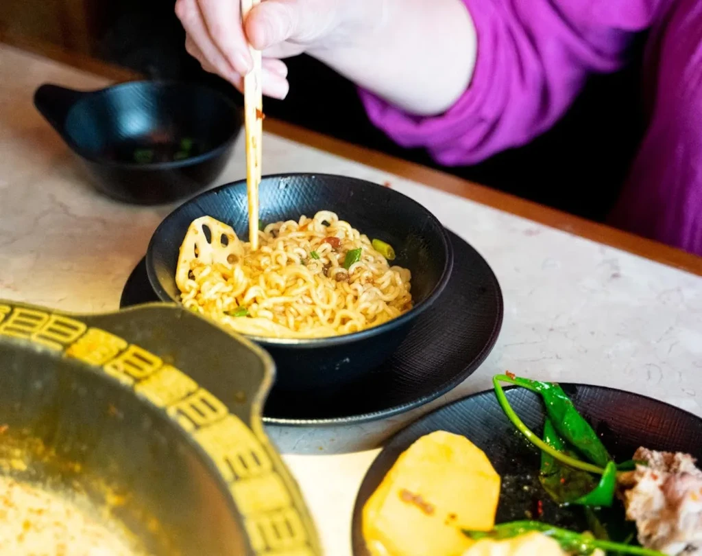 Hand with chopsticks picking yellow noodles and garnishes from a black bowl.