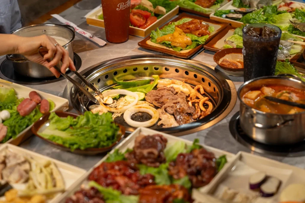 Communal grill and two hot pots surrounded by various raw ingredients.