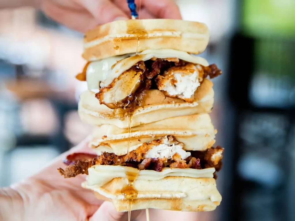 Hands hold a skewer-pierced stack of chicken, waffles, cheese, and syrup.