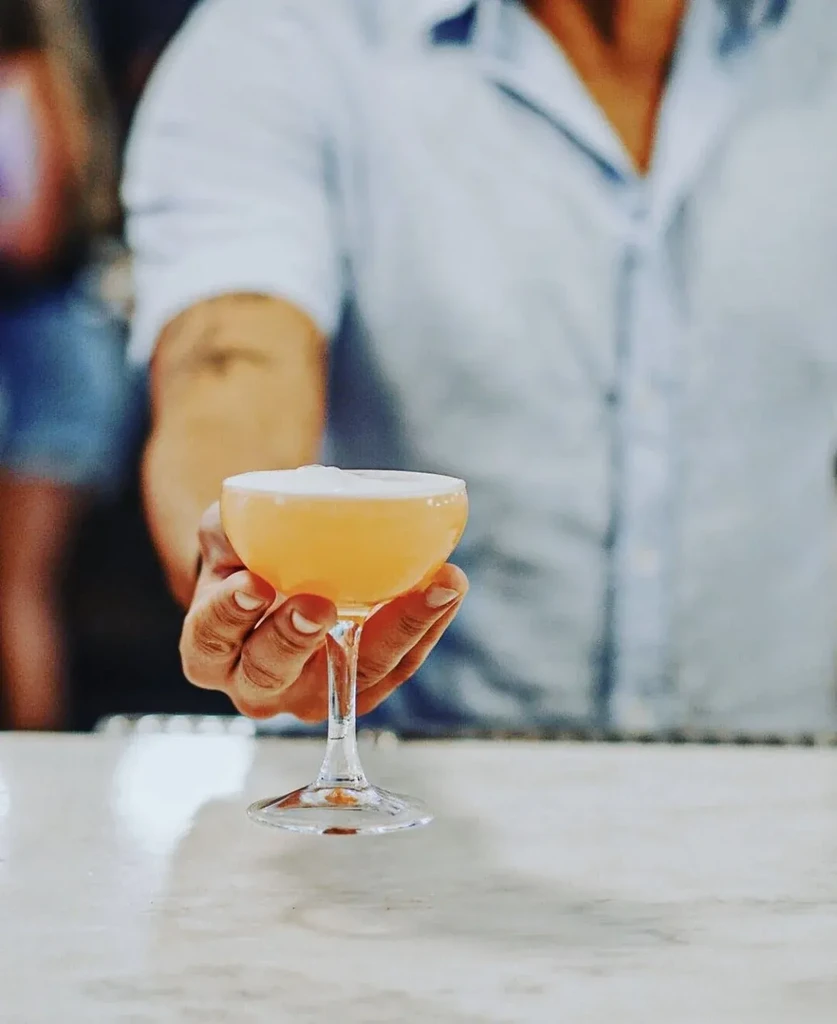 Hand holding a frothy orange cocktail in a coupe glass.