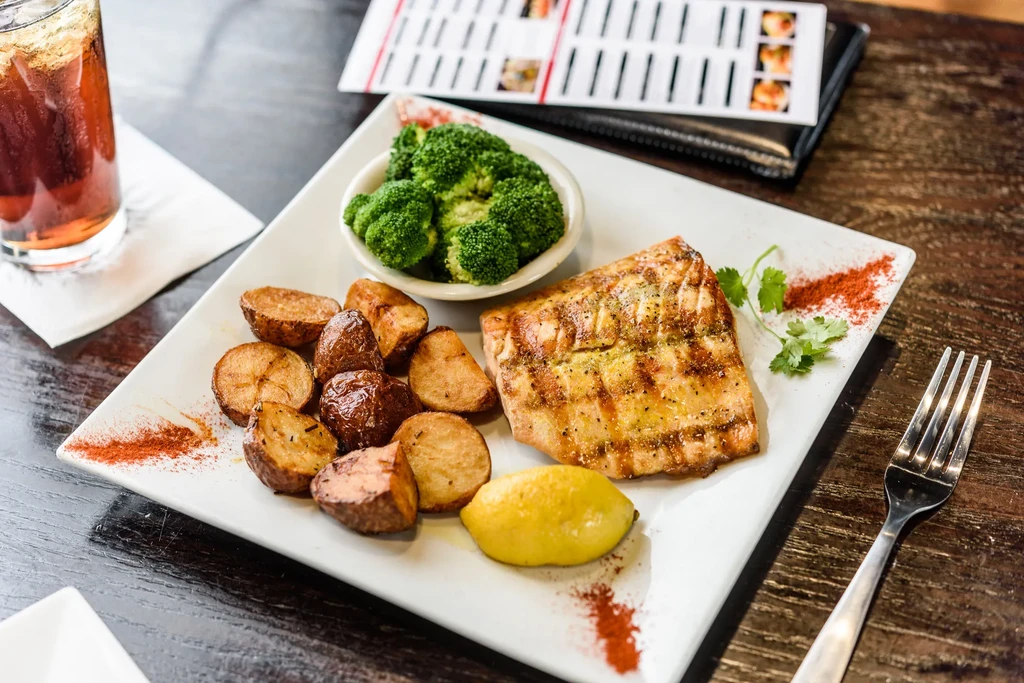 Grilled fish, roasted potatoes, broccoli, lemon, and a beverage on a plate.