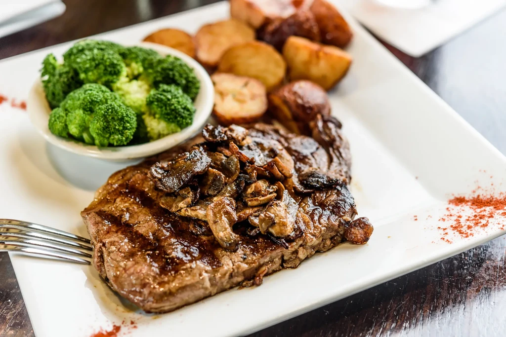 Grilled steak with mushrooms, broccoli, and roasted potatoes on a white plate.