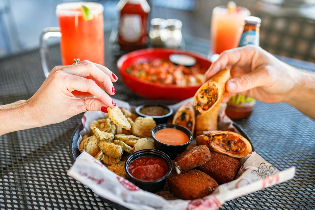 Two hands sharing fried appetizers, spring rolls, and cocktails outdoors.