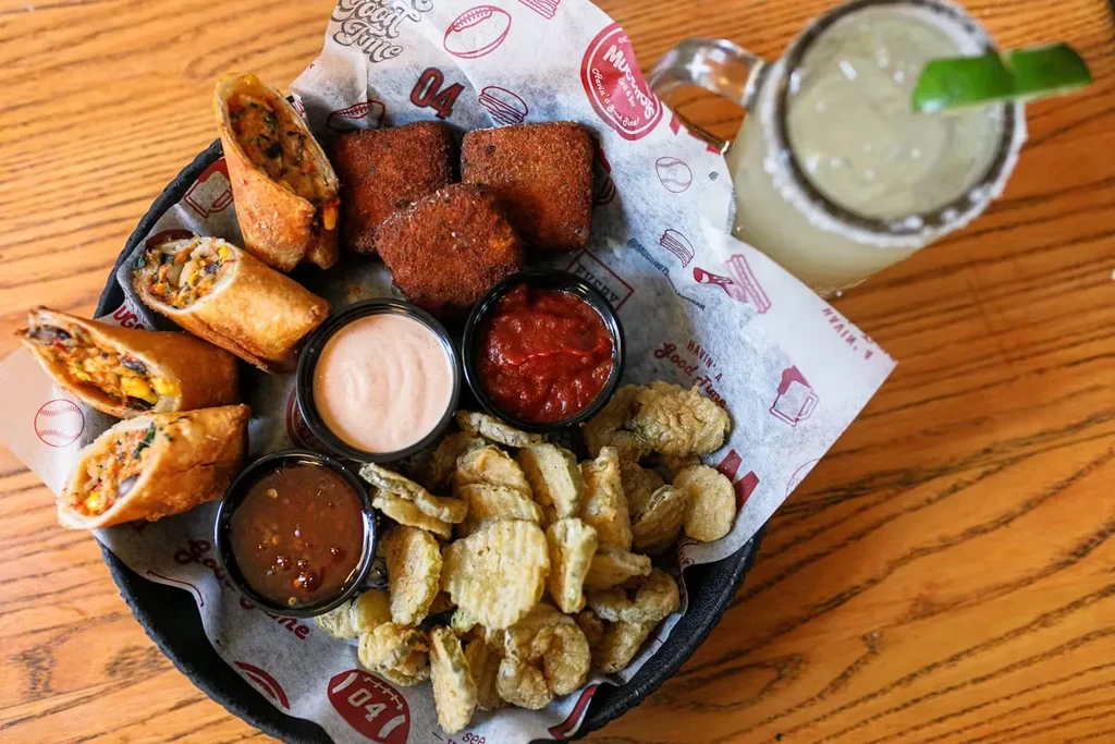 Basket of fried appetizers, sauces, and margarita on a wooden table.