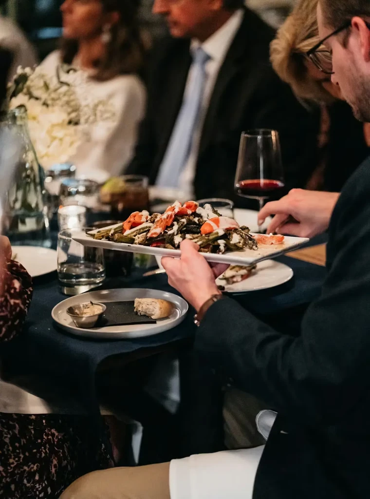Hands present a platter of grilled vegetables and carrots at an elegant table.