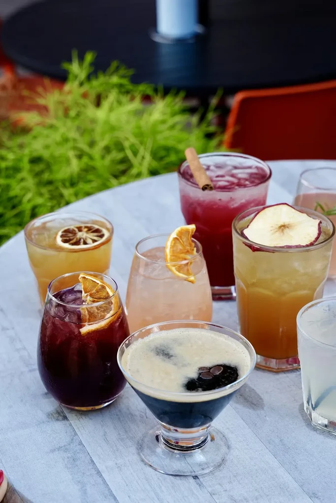 Seven colorful cocktails with garnishes on an outdoor table.