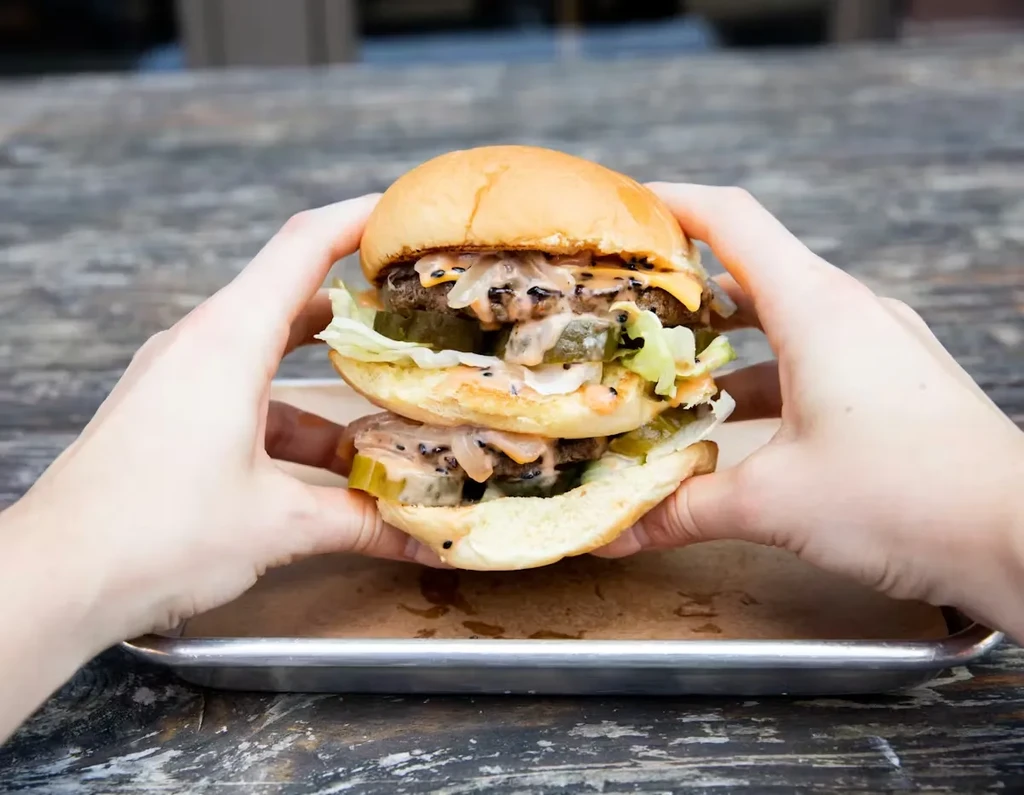 Hands holding a double-patty burger with cheese, lettuce, and pickles.