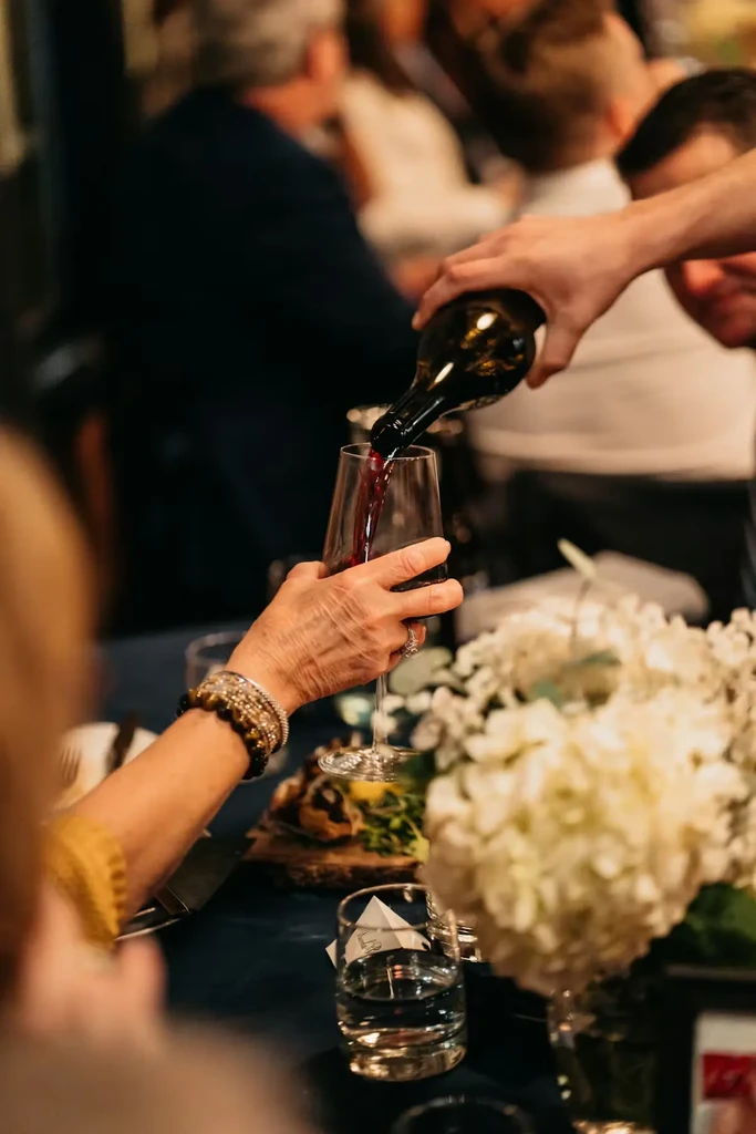 Two hands pour red wine from bottle into a stemmed glass.
