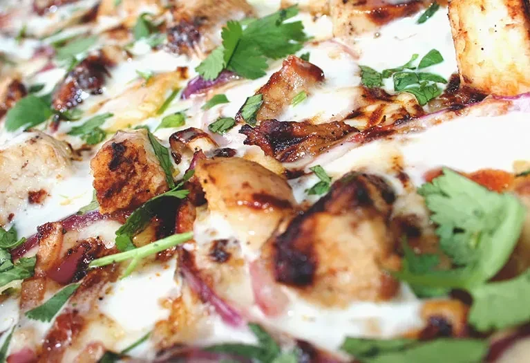 Close-up pizza with grilled meat, red onion, cilantro, white sauce, dark drizzle.