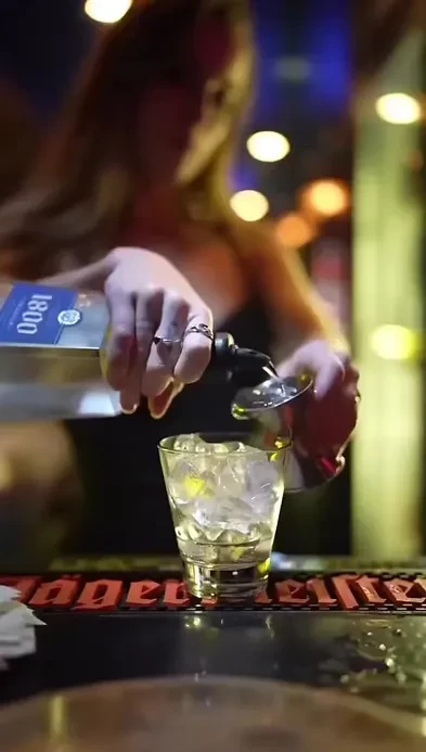 Bartender's hands pour tequila with a jigger into an iced glass.