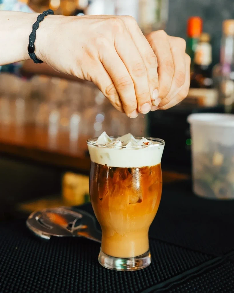 Hands sprinkle reddish powder on a layered, foamy beverage.