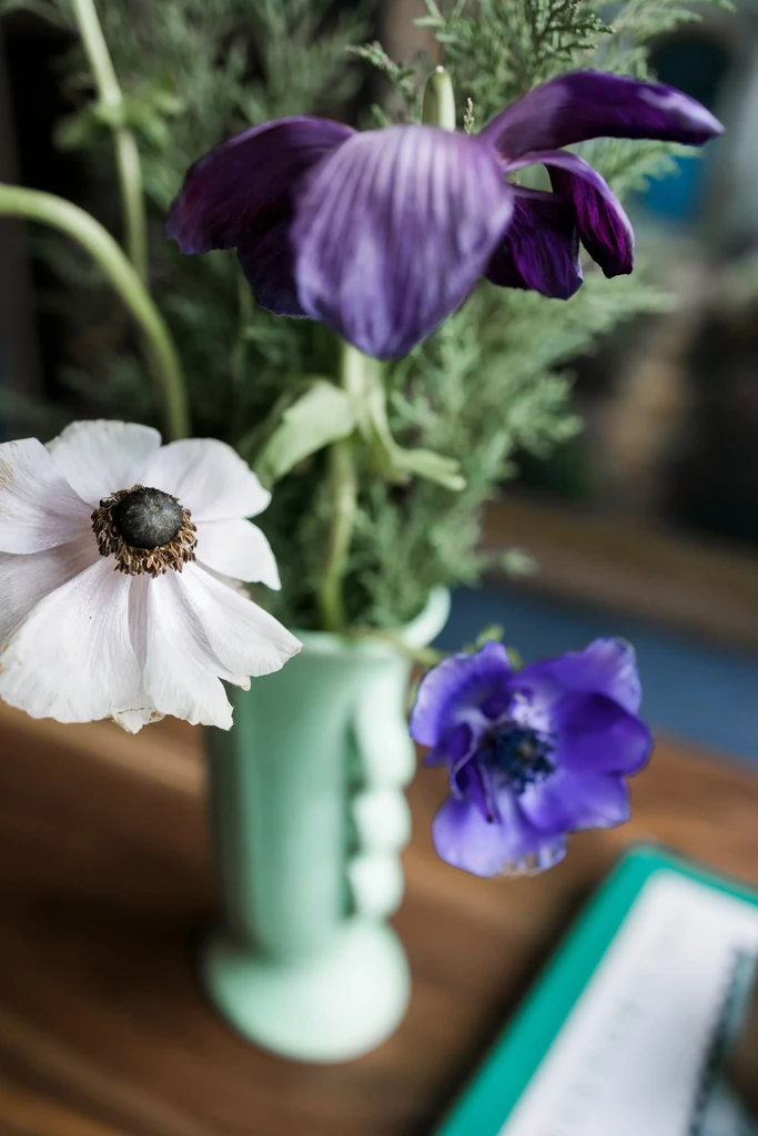 White anemone and purple flowers in a mint green vase on wood.