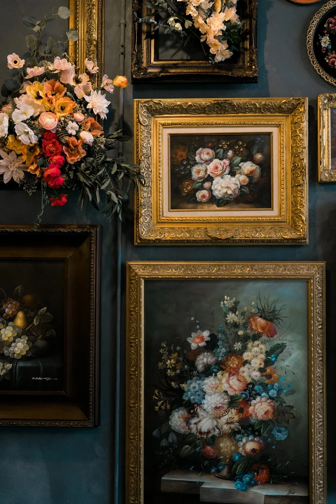 Teal wall, gilded frames, floral still-life paintings, and a large flower arrangement.